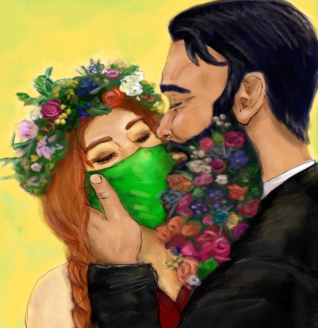 A masked woman leans into a kiss on the cheek from a dark haired man, her eyes are closed, he cups her face, she's wearing a flower crown and he has flowers woven into his beard