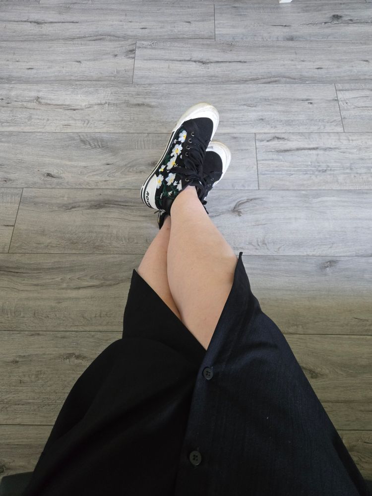 A pair of incredibly pasty legs poking out of a black dress.

The feet attached to the legs have black trainers with white flowers on them.