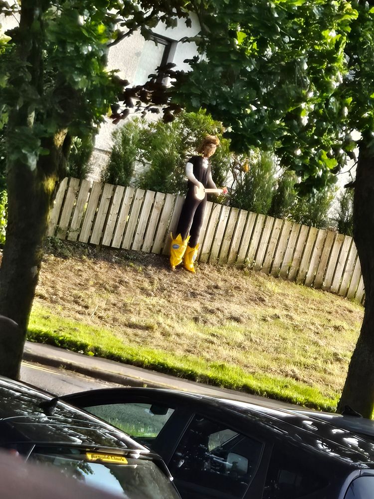 A model of Billy Connoly wrought in fabric lurking menacingly through the trees, propped up by a fence. He is wearing his iconic banana boots and holding a banjo
