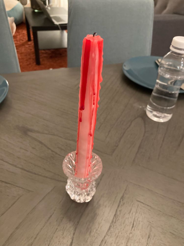 White taper candle on a dinner table. The candle is partially melted and the melted bits dripping down the sides of the candle are red, reminiscent of blood