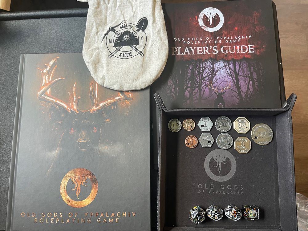 Image of the Old Gods of Appalachia Roleplaying Game rulebook, the Players Guide, the scrip coins and bag, the dice, and the dice tray. 