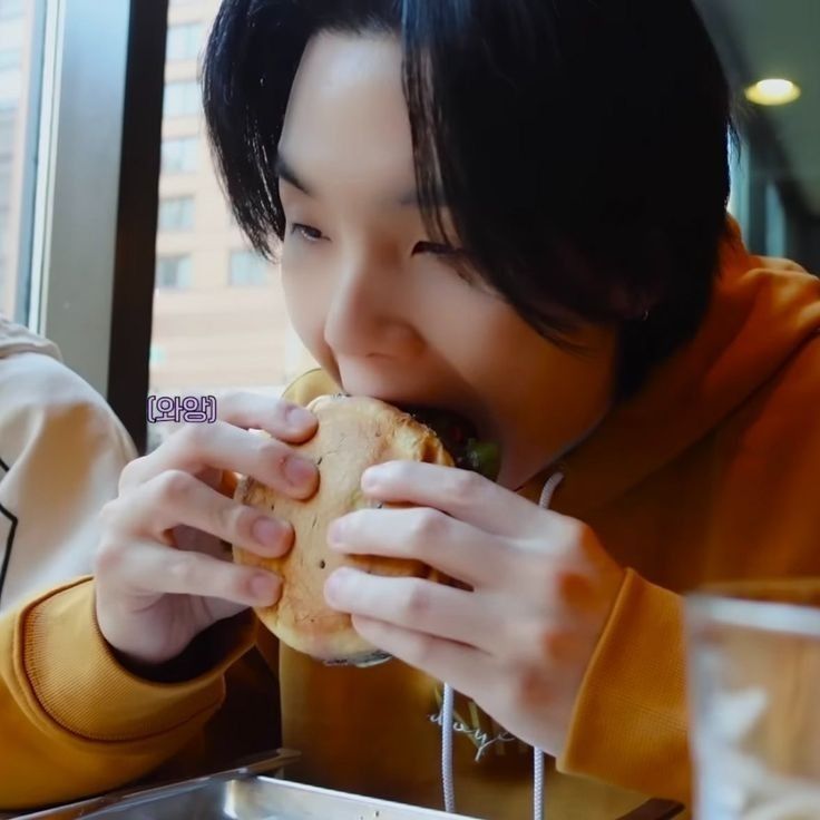 yoongi cutely taking a bite of a burger holding it with both hands. screenshot of his vlog.