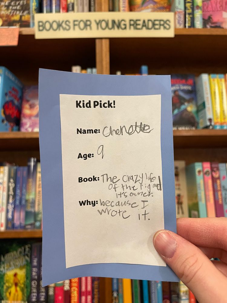 a bookmark-sized slip that says “Kid Pick!” on the top. handwritten by a child the pick says:
Name: (Illegible—Charlotte, maybe?)
Age: 9
Book: The Crazy Life of the Pig and Its Owner
Why: Because I wrote it.