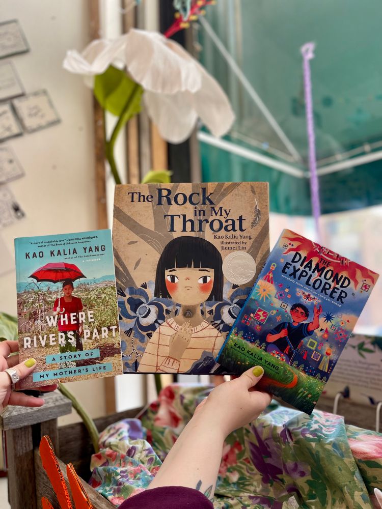 Hands holding out three books by Kao Kalia Yang, winners of the memoir, children’s, and middle grade categories: Where Rivers Part: A Story of My Mother’s Life (Memoir), The Rock In My Throat (Children’s), The Diamond Explorer (Middle Grade)
