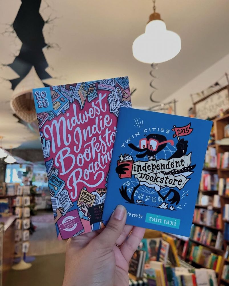 A hand holding the 2025 Midwest Indie Bookstore Roadmap and the Twin Cities Independent Bookstore Passport. Wild Rumpus’s ceiling canoe is visible in the background.
