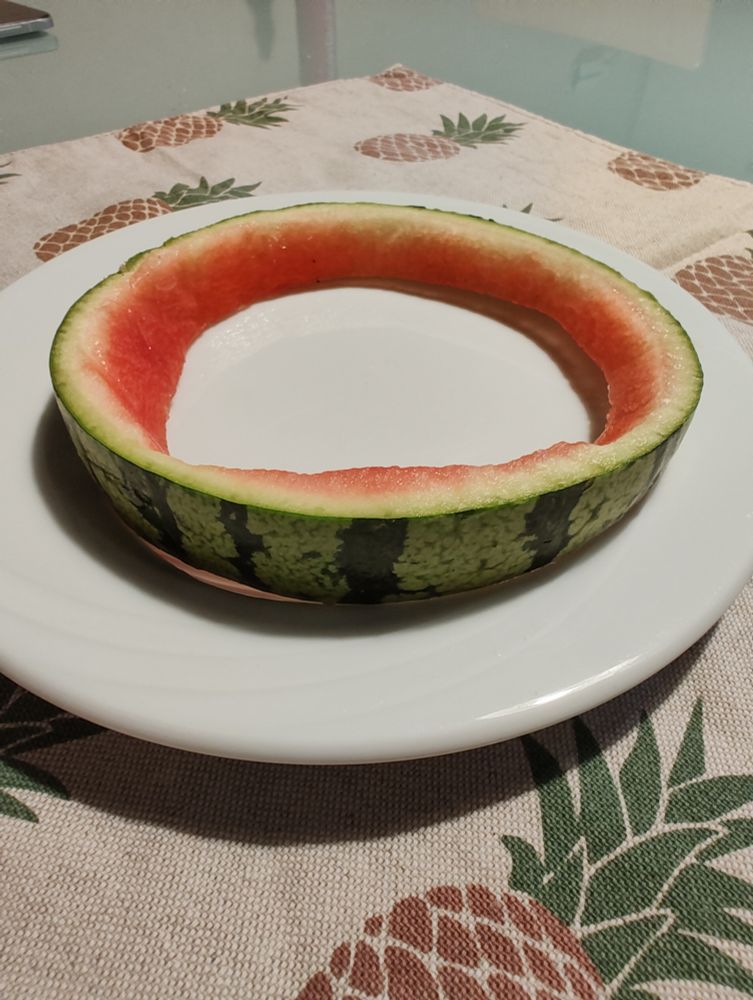 Das Foto zeigt eine halbierte Wassermelone, deren Schale dekorativ auf einem weißen Teller arrangiert wurde. Die äußere grüne Rinde mit dunklen Streifen ist gut erkennbar, während das rote Fruchtfleisch größtenteils entfernt wurde. Die Melonenschale erinnert dadurch an eine improvisierte Schüssel.

Der Teller steht auf einer auffälligen Tischdecke, die mit Ananasmotiven bedruckt ist und dem Bild eine sommerlich-tropische Note verleiht. Im Hintergrund ist ein Glastisch sichtbar, auf dem sich ein weiteres Objekt befindet – möglicherweise ein Buch oder ein Laptop –, was dem Bild eine alltagsnahe Atmosphäre verleiht.

Das Gesamtbild wirkt leicht und kreativ, ideal für einen sommerlichen Anlass oder als originelle Präsentation bei einem Picknick.