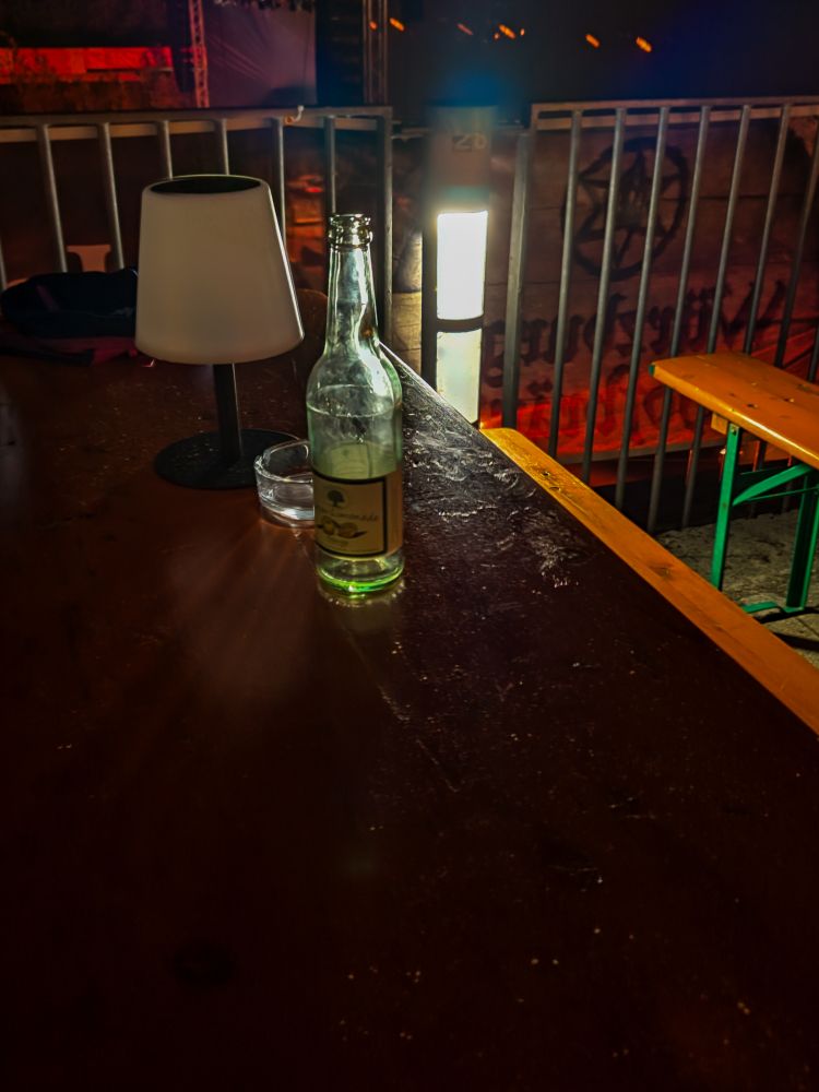 Die Aufnahme zeigt einen dunklen Holztisch im Außenbereich einer Open-Air-Veranstaltung. Auf dem Tisch stehen eine leere Glasflasche, ein Trinkglas und eine kleine Tischlampe mit weißem Schirm und schwarzem Fuß. Die Lampe ist ausgeschaltet. Rechts neben dem Tisch befindet sich eine Holzbank.

Die Umgebung ist schwach beleuchtet, mit warmen, farbigen Lichtakzenten, die vermutlich von der Veranstaltung stammen. Im Hintergrund sind Metallgeländer und eine beleuchtete Wand zu erkennen, die den Veranstaltungsbereich begrenzen. Die Szene wirkt ruhig und aufgeräumt, als wäre die Veranstaltung bereits im späteren Verlauf oder kurz vor dem Ende.