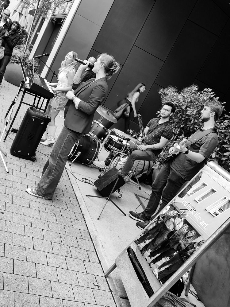 Das Foto zeigt eine Straßenszene mit einer kleinen Liveband, die im Freien musiziert. Die Aufnahme ist in Schwarz-Weiß gehalten.

Links im Bild steht eine Frau an einem E-Piano. Sie trägt ein ärmelloses Oberteil, einen kurzen Rock und hohe Stiefel. Direkt neben ihr steht eine andere Frau mit Mikrofon in der Hand, die offenbar singt. Sie trägt einen Blazer, Jeans und Turnschuhe. Dahinter sitzt ein Schlagzeuger an einem kleinen Drumset, rechts daneben ein Posaunist mit Notenständer, und ganz rechts steht ein Bassist mit E-Bass.

Die Musikerinnen sind auf einem schmalen Gehweg vor einem modernen Gebäude mit dunkler Fassade positioniert. Einige Passantinnen bleiben stehen, um zuzuhören. Auf einem Spiegel, der rechts vorne im Bild aufgestellt ist, spiegelt sich ein Teil des Publikums – mehrere Menschen, die aufmerksam der Musik lauschen.

Die Stimmung wirkt lebendig, wie bei einem spontanen Straßenkonzert, und das Schwarz-Weiß verleiht der Szene einen zeitlosen Charakter.