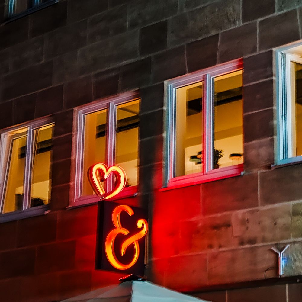 Das Bild zeigt einen Ausschnitt einer Gebäudefassade bei Nacht. Die Fassade ist dunkel, aber mehrere Fenster sind von innen beleuchtet, was auf eine belebte oder genutzte Umgebung hinweist. Besonders auffällig ist ein leuchtendes Neonzeichen an der Wand: Es zeigt ein rotes Herzsymbol und ein „&“-Zeichen. Dieses Neonlicht strahlt hell und hebt sich deutlich von der dunklen Umgebung ab. Es wirkt wie ein dekoratives Element, das Aufmerksamkeit erzeugt und möglicherweise auf ein Geschäft, eine Bar oder ein kulturelles Angebot hinweist.

Die Gesamtstimmung des Bildes ist urban und ruhig, mit einem klaren Fokus auf das leuchtende Symbol in der Dunkelheit.