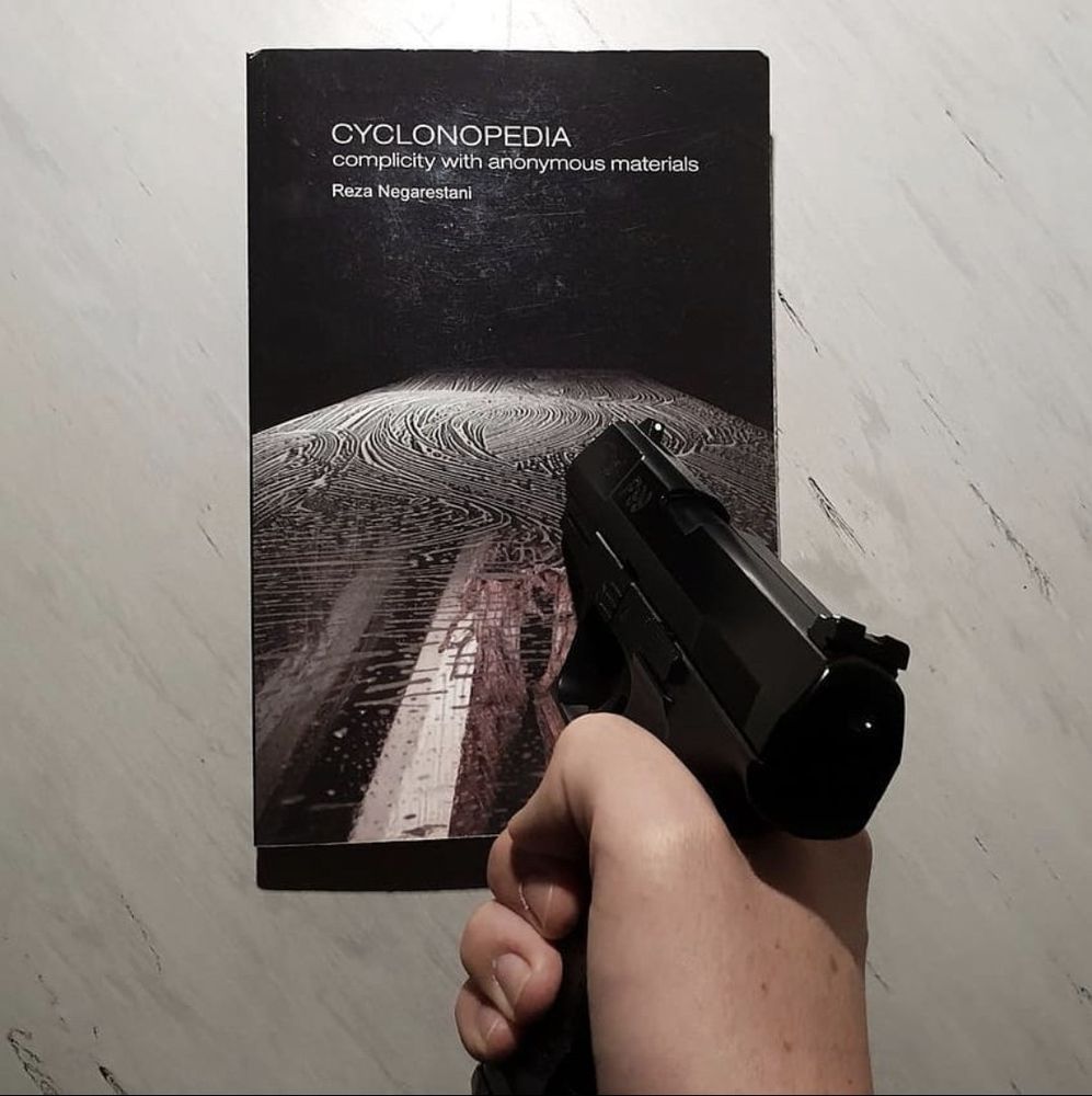 A hand angrily pointing a pistol at Reza Negarestani’s “Cyclonopedia: Complicity with Anonymous Materials,” a work of theory-fiction which posits that oil is a malevolent, sapient, eldritch entity trying to destroy the earth and turn mankind against the sun. 