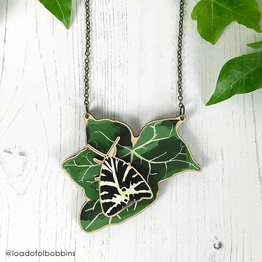 A laser cut wooden statement necklace by Loadofolbobbins. It shows an illustration of lush dark green veined ivy leaf with a Jersey tiger moth perched atop it. The jersey tiger moth stands proud of the leaf. The moth has black and yellowy cream zebra like stripes on it's wings, which are tucked behind it looking like an arrow head shape. The necklace is hung from a antique bronze cable chain. The necklace is laying against a white wood background and surrounded by real ivy clippings.