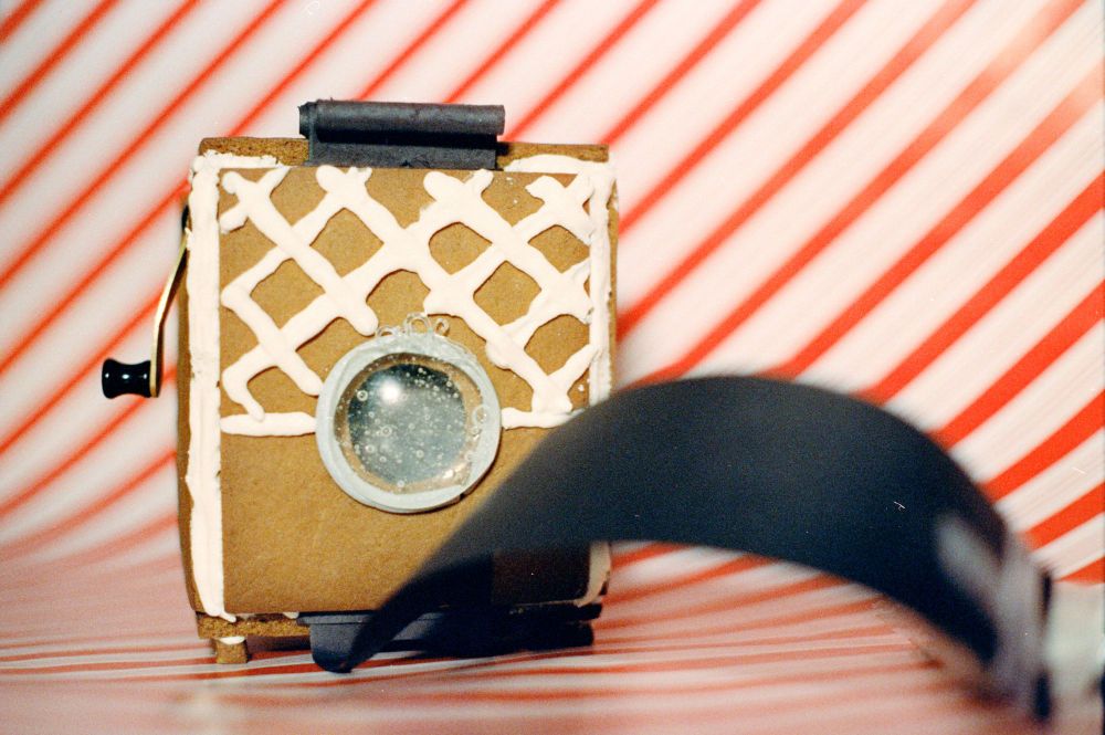 A boxy film camera made of gingerbread with a large sugar lens and a paper shutter haning from the top and bottom. It has a criss-corss icing sugar pattern on the top half. The camera ion a white/red stripe wrapping paper background