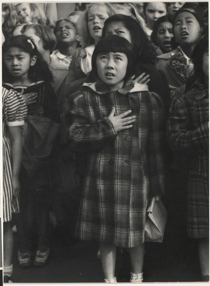 A young Japanese girl stands front and center amid a crowd of young girls. Her hand is on her chest as the girls recite the pledge of allegiance. Her eyes, which wear a look of distress and uncertainty, point upwards towards a flag hidden somewhere behind the cameras