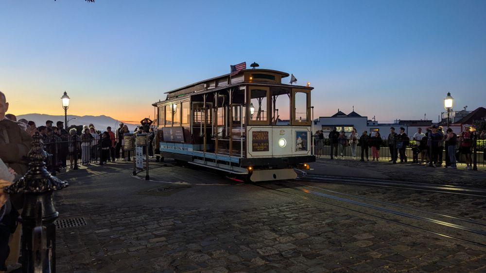 A San Francisco trolley car being rotated on the turntable. The sun is setting. The hills and Golden Gate bridge is lit dimly in the background. There is a long line of people surrounding the trolley.