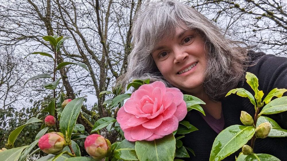 A smiling middle-aged woman behind a pink flowered Camellia japonica 'Mrs. Tingley'.  Bare trees, and grey skies, are in the background.  The woman has graying brown hair and is wearing a black jacket.