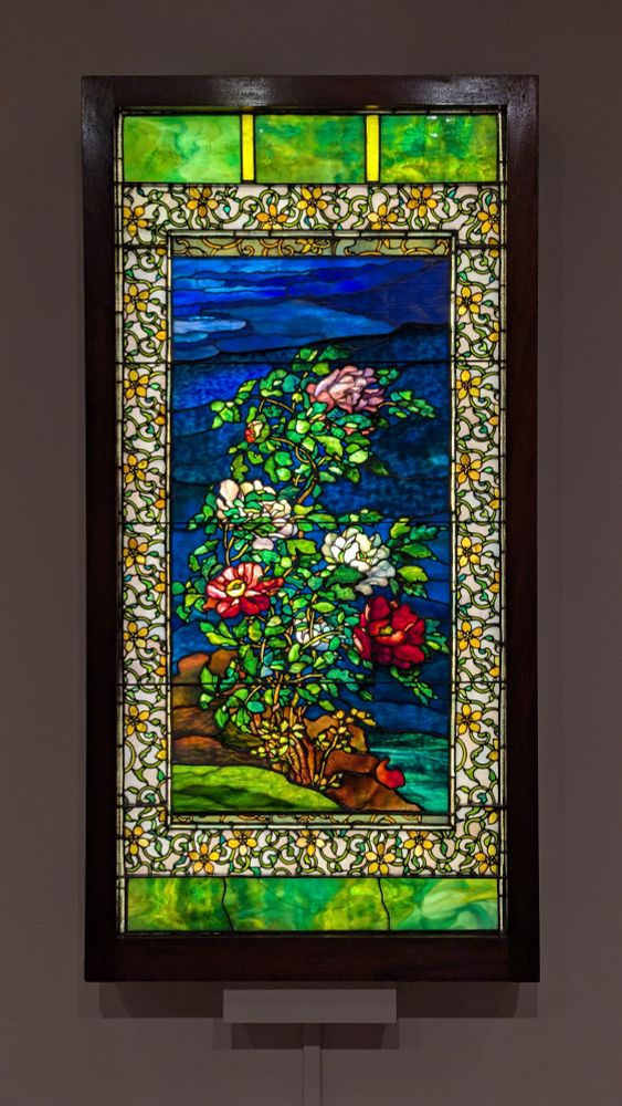 A rectangular stained glass window that has been converted to a light box. The stained glass peonies are pink, white, and red, with green and chartreuse leaves. The image is set on a blue backdrop. The main image is surrounded by a colorful floral frame. The top and bottom edges are lime green. The wood frame is finished in black.