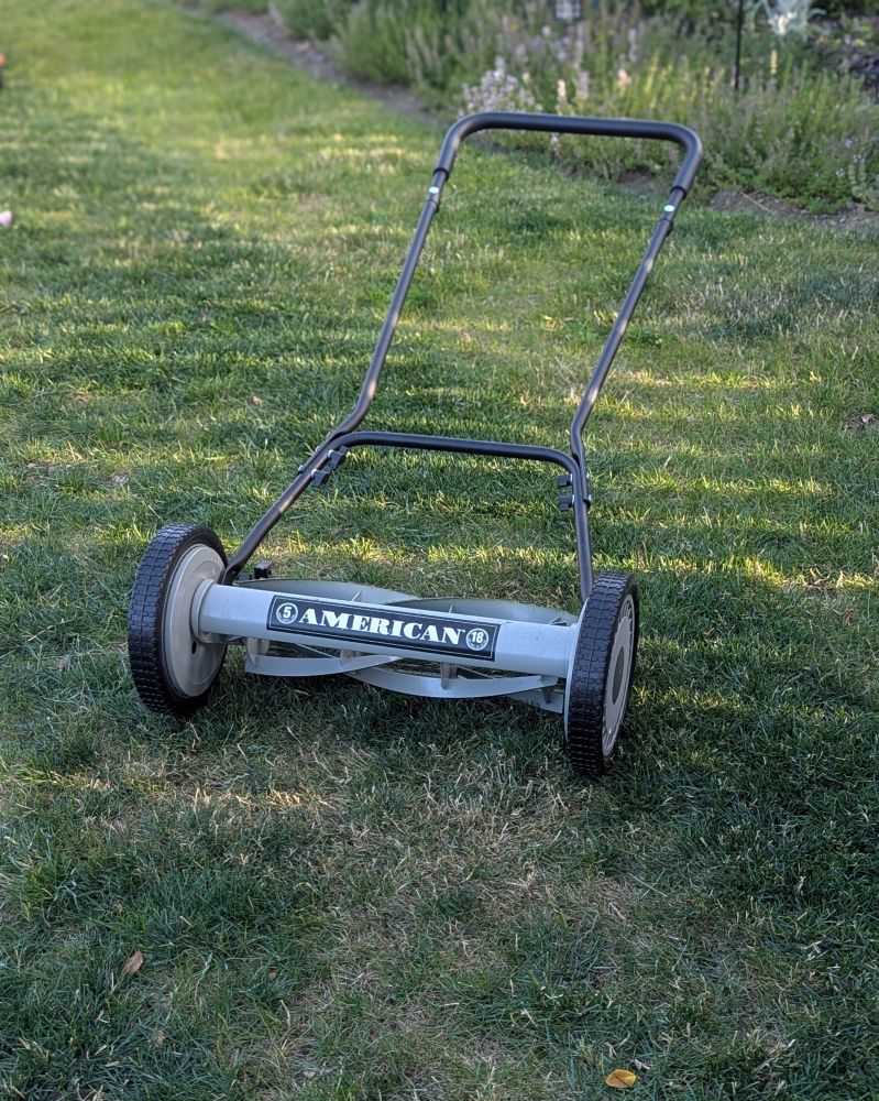An old fashioned style push mower that was manufactured by the American Lawn Mower Company in Indiana. The lawn mower is grey, with black wheels and a black handle. The mower is sitting on freshly mowed grass that is browning due to the high heat of summer.