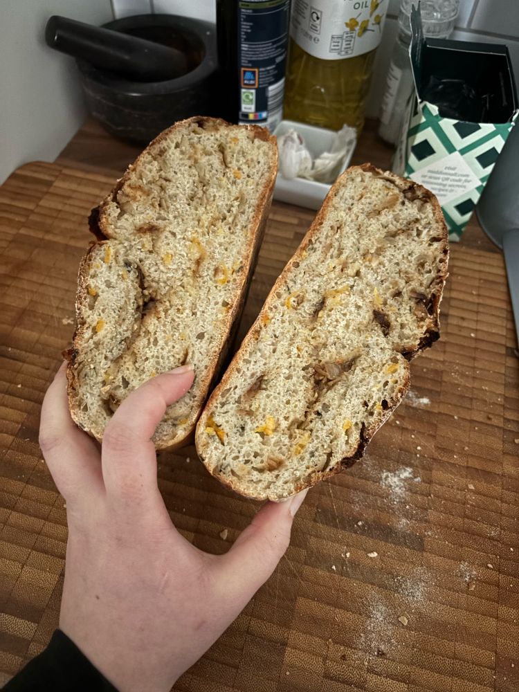 A large loaf of bread, cut in half to show a cross section, on a wooden chopping board. The bread has dark brown swirls and little orange bits - shallots and apricot. 
