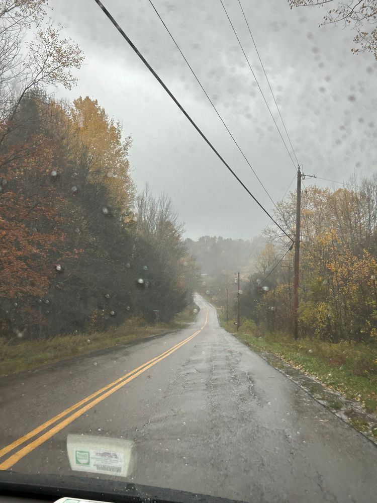Much needed rain after a long drought in Vermont, the rain clings to the windshield as the viewer drives down a winding mountain road. The road is framed by trees in muted fall colors.