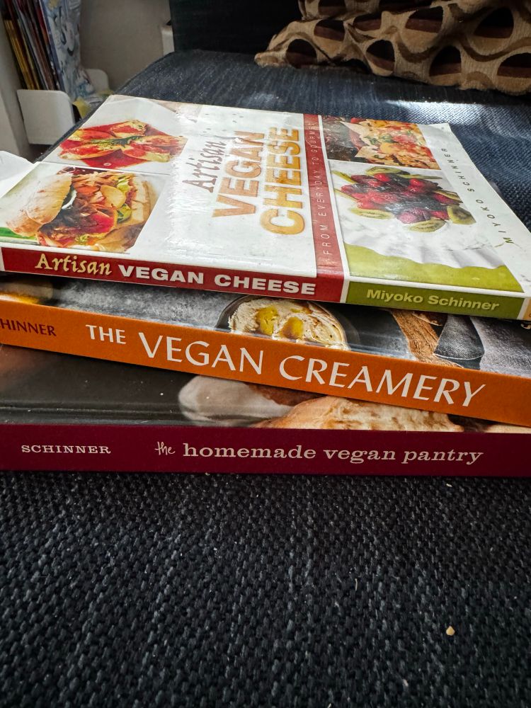 A stack of three vegan cookbooks on a blue couch, Artisan Vegan Cheese, The Vegan Creamery, and Homemade Vegan Pantry all by Miyoko Schinner 