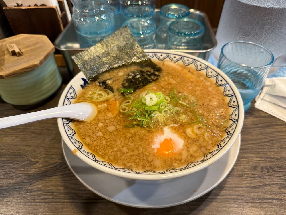 ラーメンの写真。ばんから池袋東口店様にて。背脂がたっぷり浮いた茶色のスープ、温泉卵が浮かび、ネギと海苔がトッピングされている。