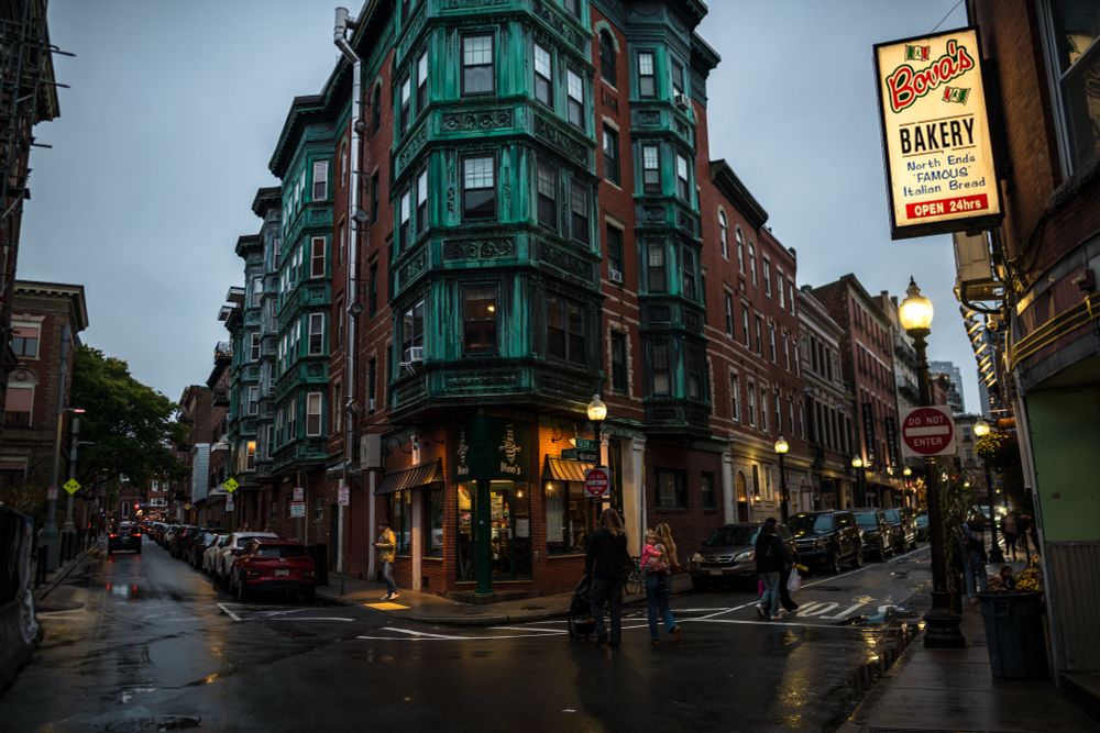 A wet North End, Boston intersection at dusk. Golden street lights light up the divergent intersection. A triangular brick building with copper bay windows anchors the intersection.