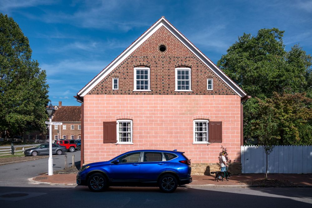 A man walks his dog past a a blue SUV in front of a Colonial-era building in Wintson-Salem, North Carolina. The symmetrical facade with four offset cross-hatched windows features a pink stone facade with a brick peak.