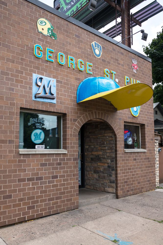 A giant baseball cap protruding from a simple brick facade with the logos of most of Wisconsin's sports franchises. 
