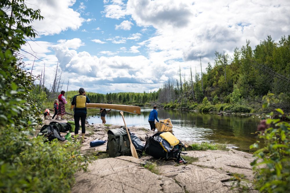 Portaging from Alpine into Jasper Lake. The kids and wife wait while their uncles place a pair of Kevlar canoes into the water from a rocky shore. The young forest is a radiant green along the shores.