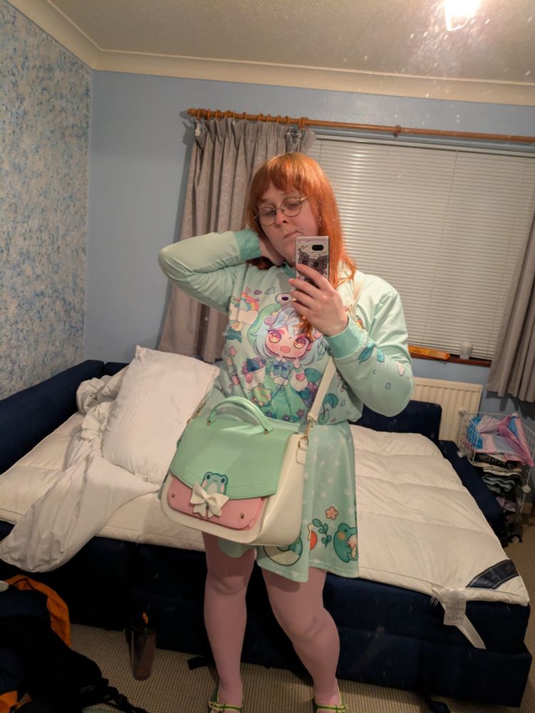 A woman with ginger hair taking a selfie, wearing a froggy skirt, a jumper with an anime girl wearing a frog dress, and a froggy bag