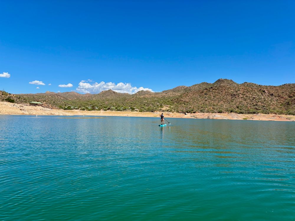 A person paddles on a stand-up paddle board in a serene lake surrounded by desert hills