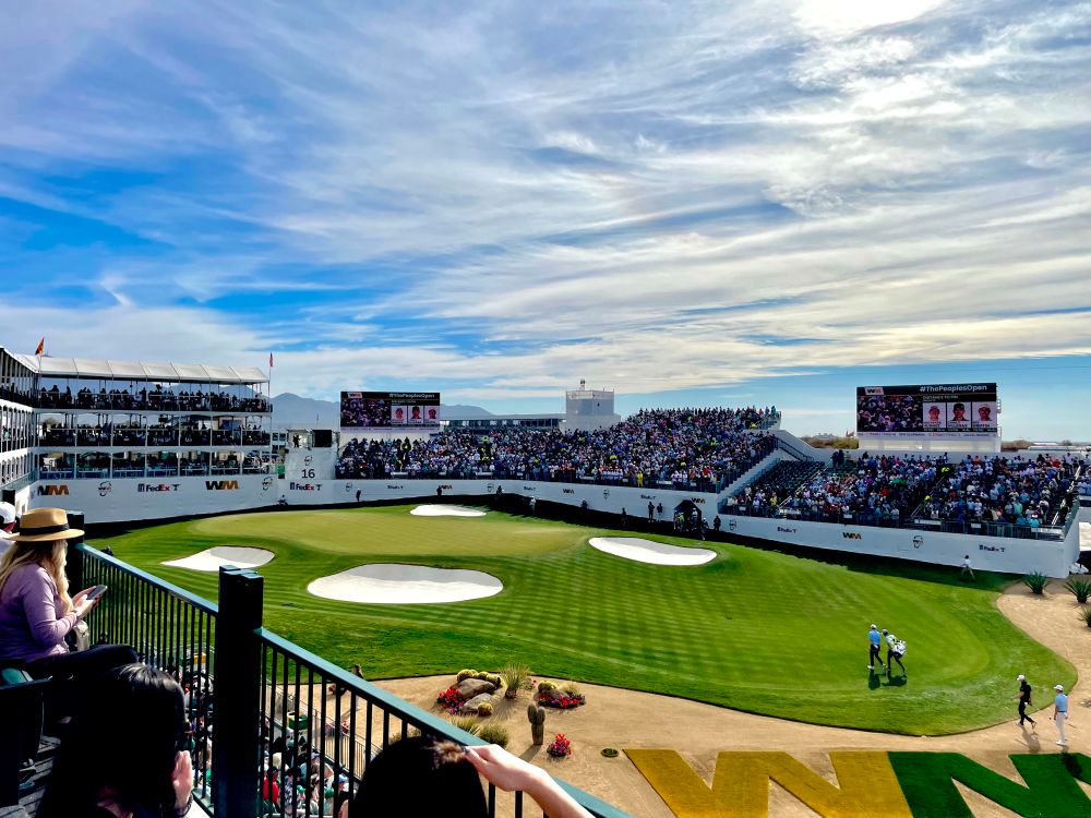 Golf tournament at crowded stadium, with fairway, sand traps, grandstands, and branded banners under a blue and cloudy sky.