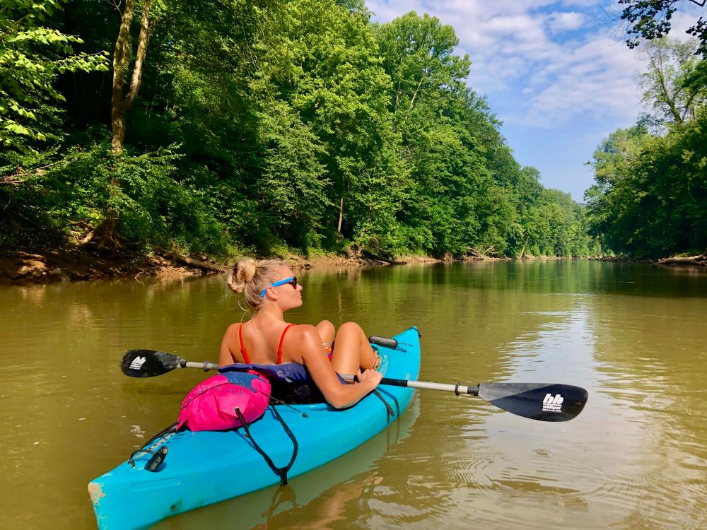Woman kayaking on a river, surrounded by green trees under a partly cloudy sky.