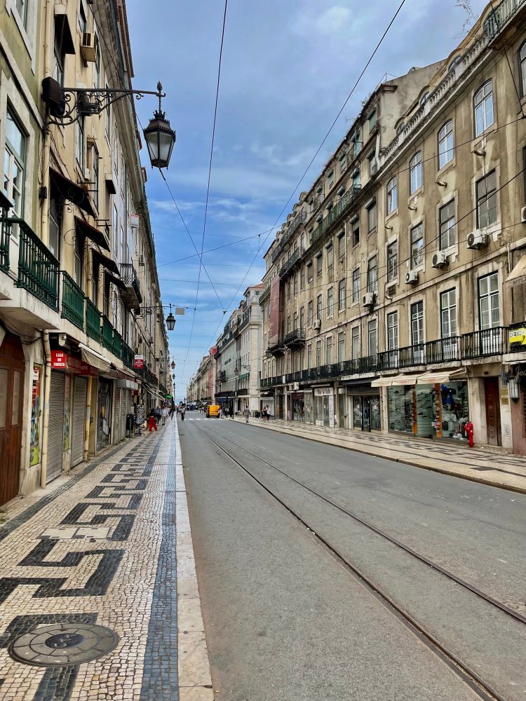 A street in Lisbon, Portugal, showcasing aged, multi-story buildings lining each side. The street features tram tracks running down the center. The sidewalks are paved with a distinctive black-and-white geometric pattern. Overhead wires crisscross the sky, and historic street lamps adorn the buildings. A few pedestrians and a yellow vehicle are visible in the distance.