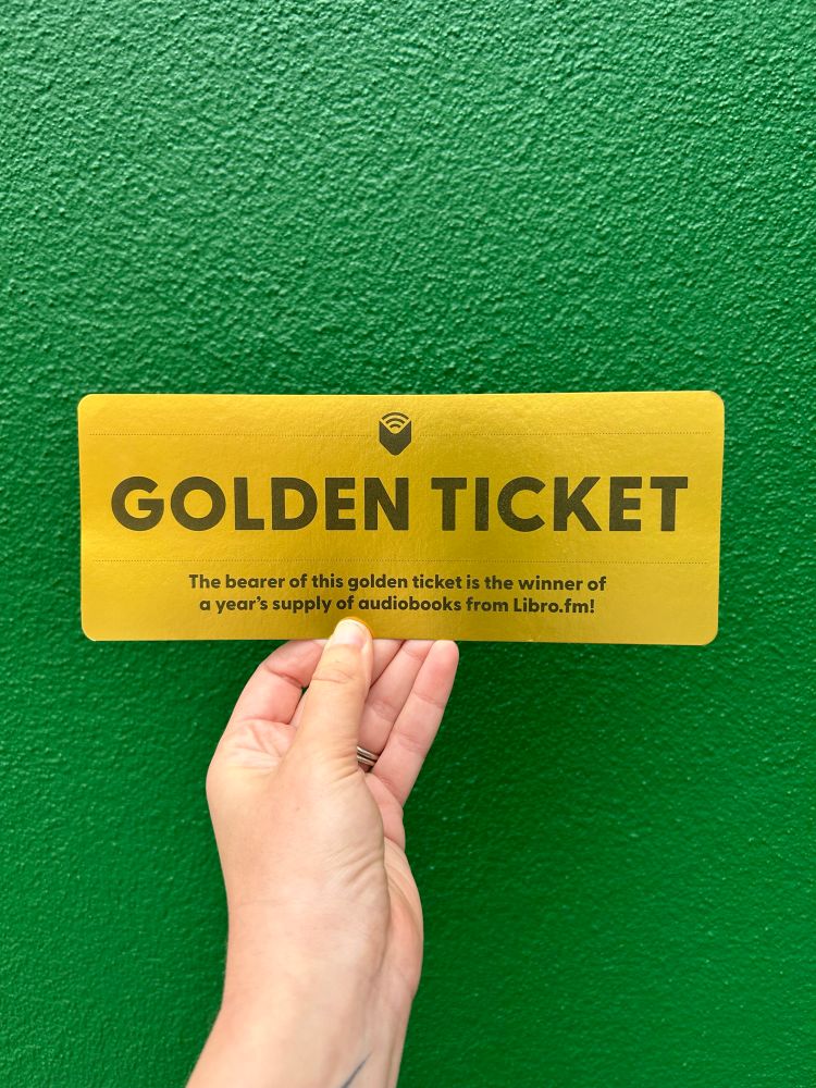 Photo of a hand holding a Golden Ticket against a green wall. The ticket reads “golden ticket, the bearer of this golden ticket is the winner of a year’s supply of audiobooks from Libro.fm”