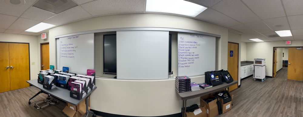 Panorama photo of a room with textbooks organized by publisher and whiteboards with table of contents written. 