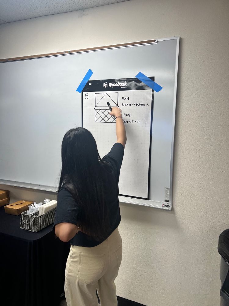 Teacher writing on a wipebook with grids drawn to show a billiard ball path along 45 degree angles with different dimensions of a table.