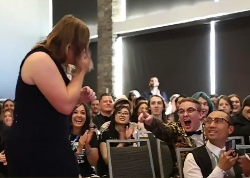 screenshot of frootoon about to walk to the stage to receive her award, hand over her mouth in disbelief. she is looking at sylleblossom, who is enthusiastically pointing at her and shouting "that's my girl!"