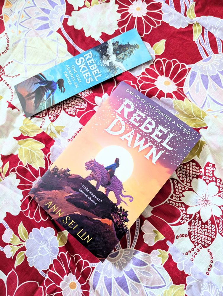 Rebel Dawn book cover features a girl riding an origami tiger as the sun rises behind her. The book lies on a red and white flower patterned yukata, next to a blue bookmark 
