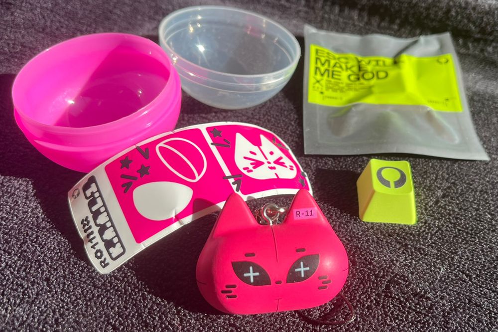 The gachapon's contents. The pink capsule contained a sticker with the face of KAAL1, a robotic cat. A keychain also depicts KAAL1's face. Next to it, a green keycap bearing the Marathon logo (an incomplete circle, with a slice down the bottom middle). Packaging is visible for the keycap, which reads ESCAPE WILL MAKE ME GOD.