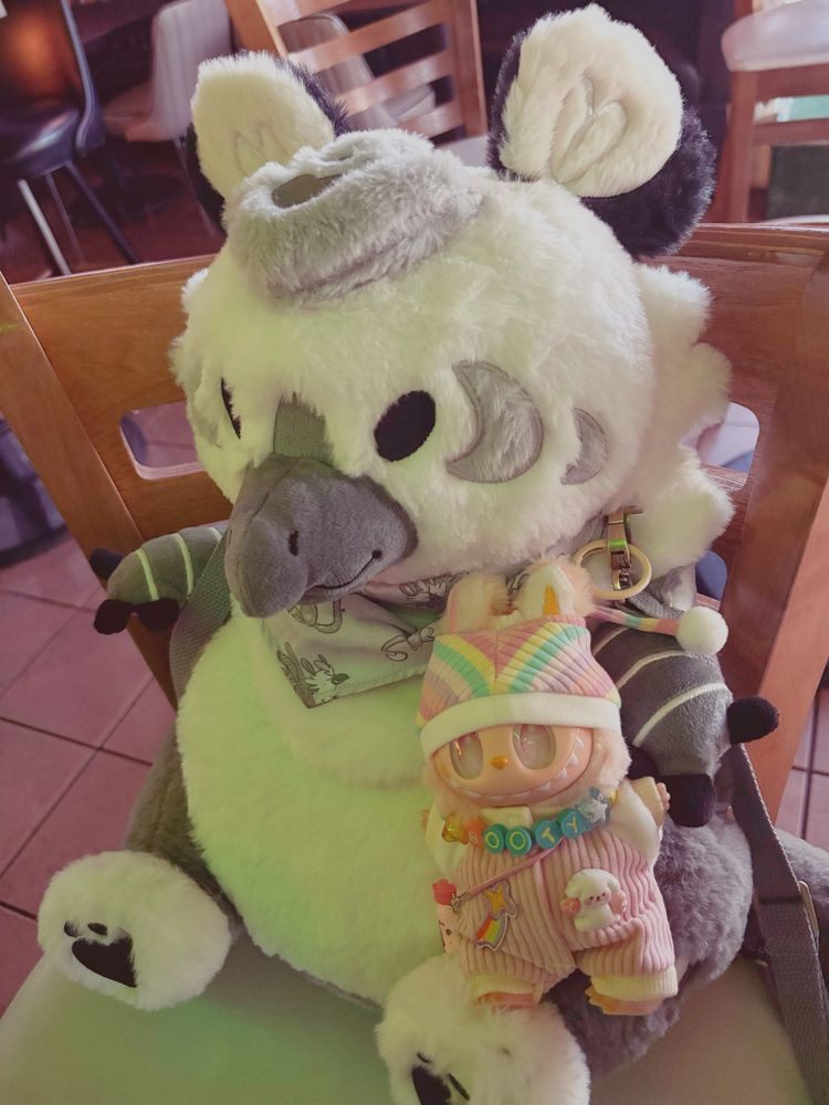 A bootleg peach Labubu wearing a rainbow hat, pink jumpsuit with a rainbow shooting star and a white puppy charm, carrying a pink milk bottle on a strap, and wearing a necklace that says "booty," is posed sitting on the leg of a giant plush gryphon backpack. The gryphon is silver and white, chubby, and has gray straps. The two are sitting on a chair at a table in an Irish pub.