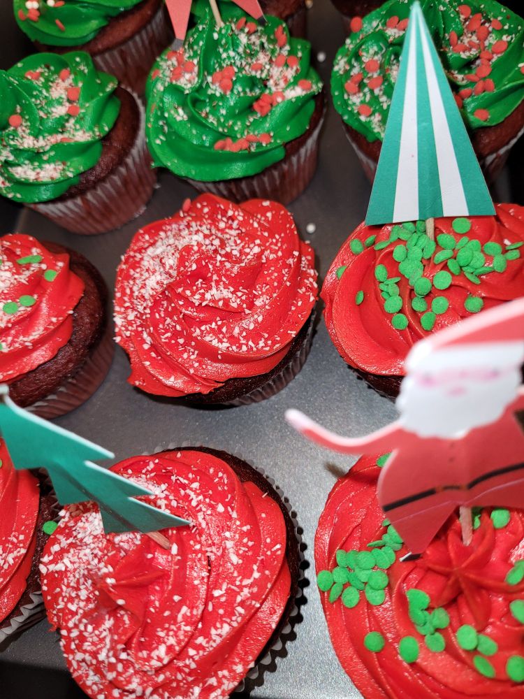 Red velvet cupcakes with red and green frosting. Some have green or red sprinkles and some have peppermint dust. Some have paper reindeer, Santa or Christmas tree paper toppers. 