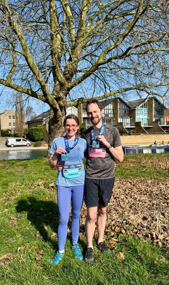 Rebecca and Robert holding medals in athletic clothing standing in a field in front of a tree and a river 