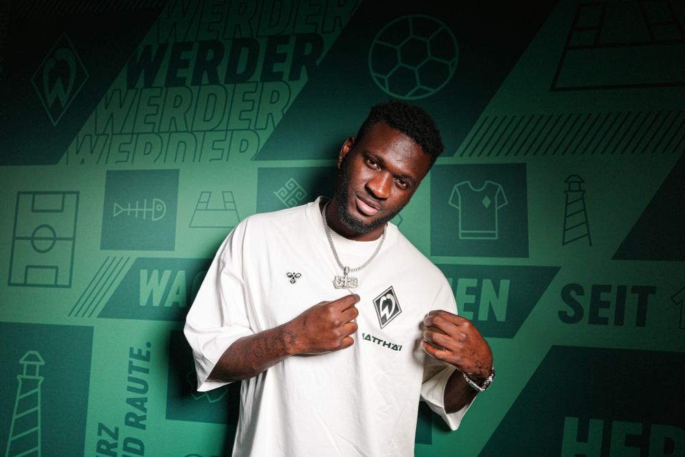 Victor Boniface steht im einem weißen Trikot vor grünem Grund. Er hält mit seinen Händen die Werder-Raute.