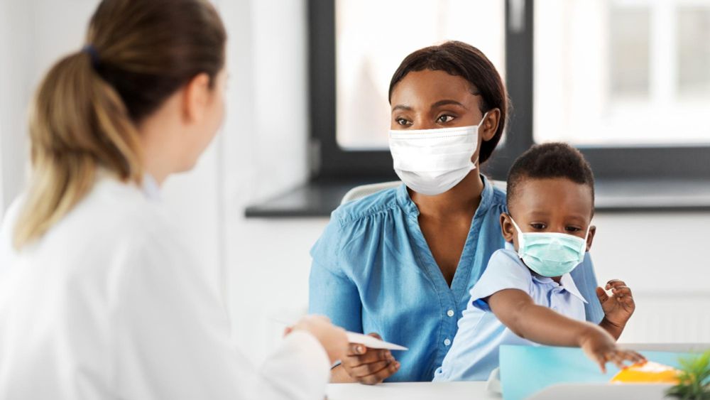 A person wearing a mask sits at a table with a young child on their lap, also in a mask, during a consultation with a healthcare professional in a medical office.