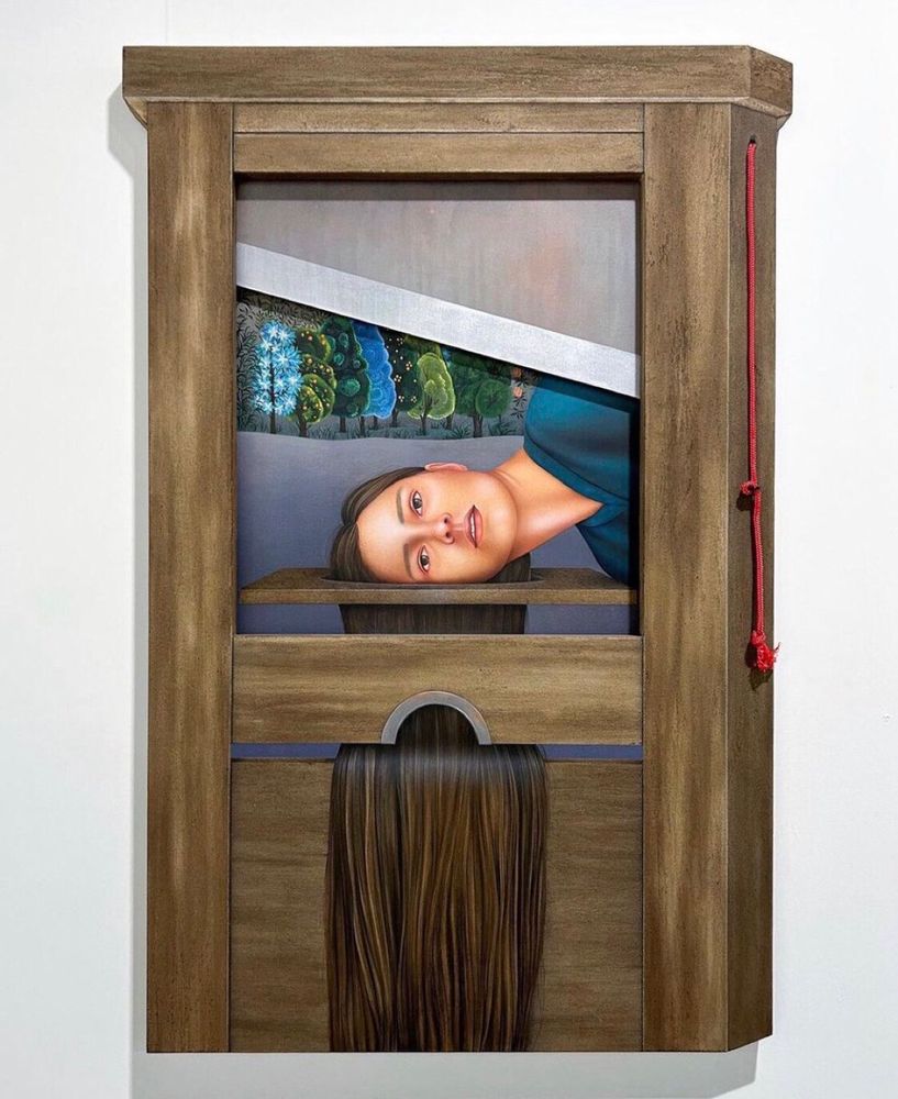 Installation, die aussieht wie ein Fallbeil, dahinter ist das Gesicht einer jungen Frau zu sehen. Ihre langen braunen Haare liegen unter dem Fallbeil. Im Hintergrund Bäume.