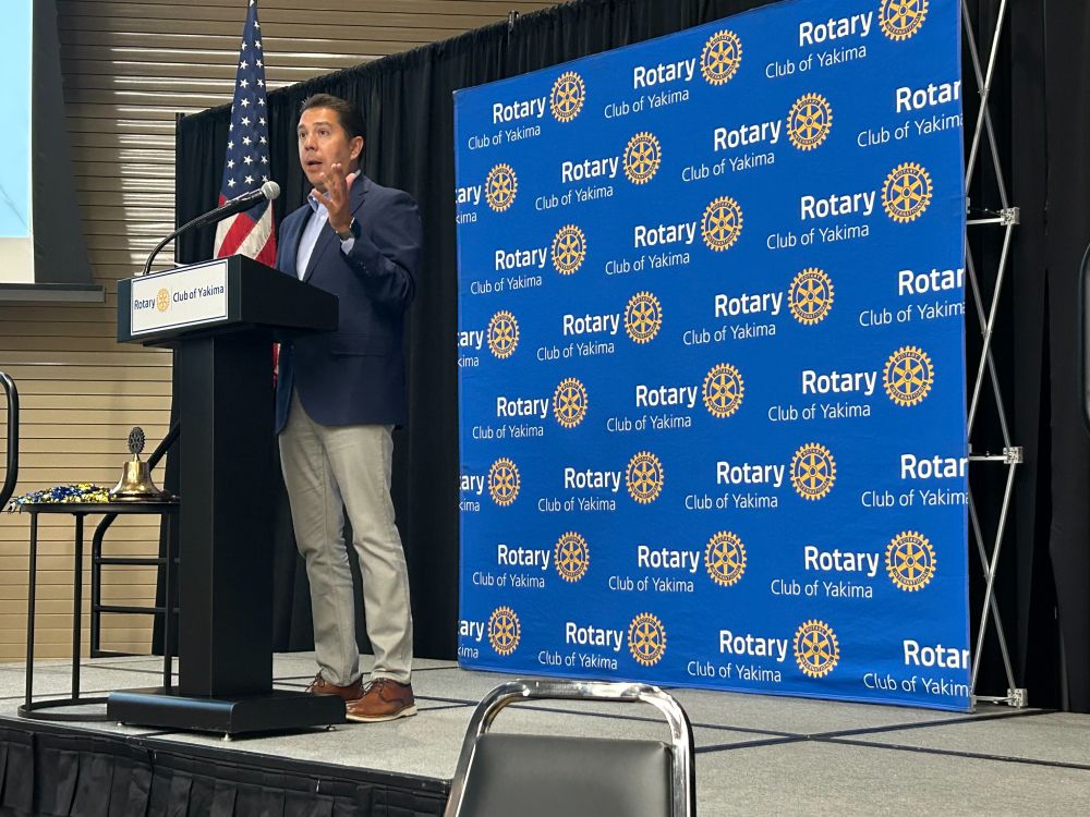 A person in a blue jacket stands speaking behind a black podium on stage. The podium has a white sign reading “Rotary Club of Yakima,” and at the back of the stage is a blue backdrop reading “Rotary Club of Yakima.”