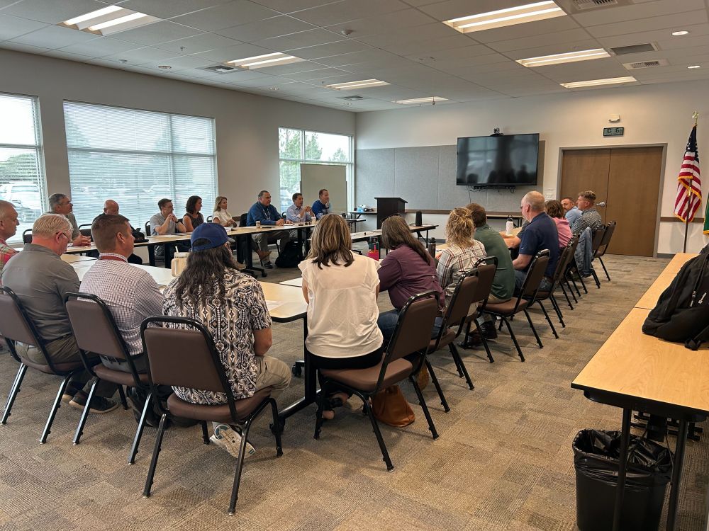 A large group of people is seated at long tables lining a conference room. They are looking at one person speaking, all seated in brown chairs with large windows on one wall.