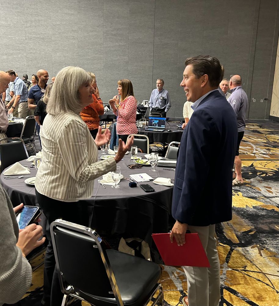 A person in a white and gray striped shirt gestures while speaking to a person in a dark blue jacket. They are standing in a room full of people at circular black tables.