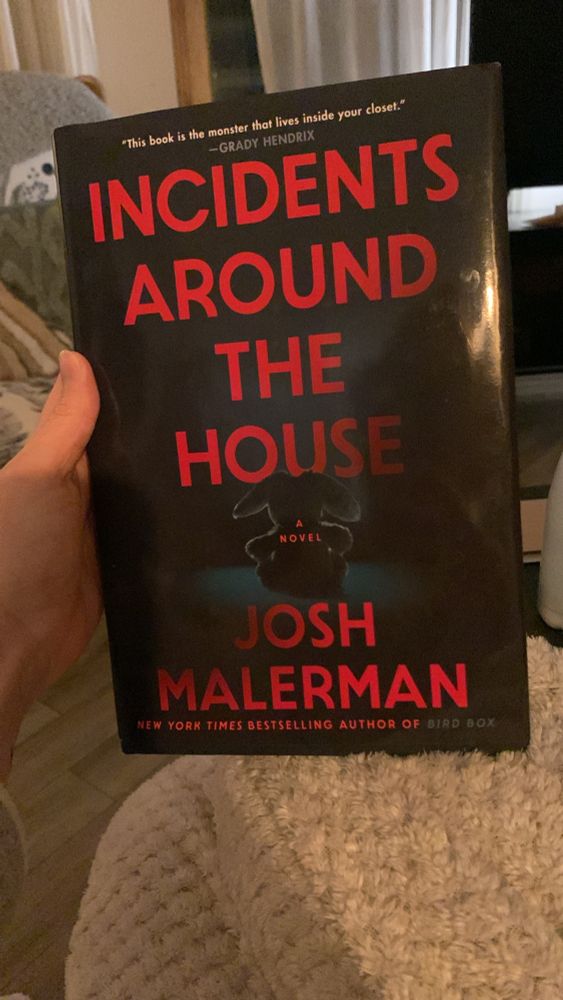 A hand holds up a novel with a black cover and red lettering for the title, “Incidents Around the House by Josh Malerman”